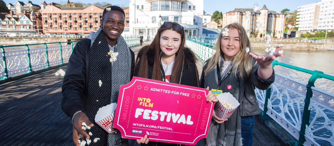 3 young people stand on Penarth Pier throwing popcorn in the air and holding a giant cinema ticket that reads 