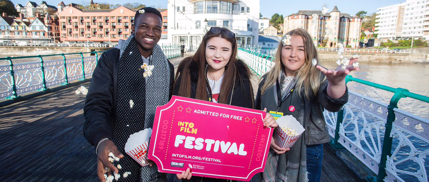 3 young people stand on Penarth Pier throwing popcorn in the air and holding a giant cinema ticket that reads 
