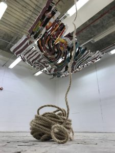 Art work is suspended from a warehouse ceiling of corrugated metal. On the floor a large swirl of thick rope stretches up to the ceiling.