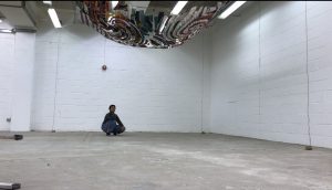 Jodi squats on a concrete floor below her artwork Benjamin which is suspended on the ceiling below fluorescent tube lighting. The image is dominated by the white brick walls and stark flooring. Jodi looks small as she is at a distance. 