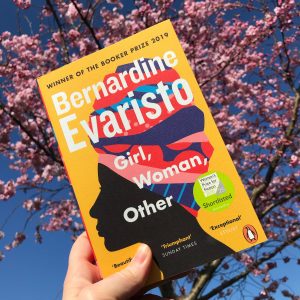 The book of Girl, Woman, Other by Bernadine Evaristo is held by hand against a background of a pink blossoming tree in front of a bright, clear blue sky. The book cover is a strong yellow with a silhouette of a black woman with a multi coloured headscarf. 