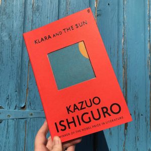 The book of Klara and the Sun by Kazuo Ishiguro is held up by hand in front of a painted blue wooden background in the sun. The book cover is a striking red colour with bold black text and a square central image of the sun just peaking into a blue sky. 