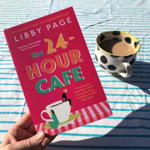 The book of The 24 Hour Cafe by Libby Page is held by hand at a jaunty angle with a background of a striped tablecloth and a spotty mug of tea. The book is a bold pink colour with an illustration of a cup and saucer with two female characters sitting on it like Borrowers.