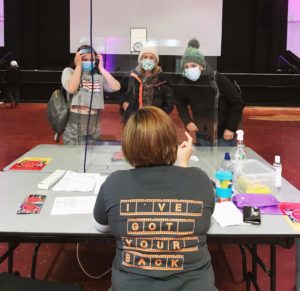 A volunteer sits centrally in the foreground with her back to the camera. On it, the t-shirt reads I've got your back. Three participants in masks are listening to her give directions. They are in the Memo's main space.