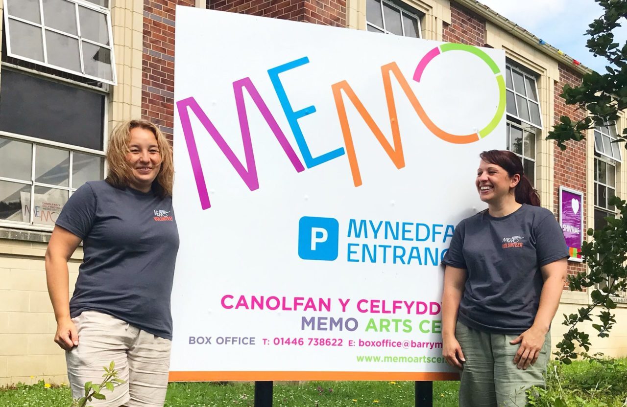 two volunteers stand either side of the large Memo sign outside the building. They are wearing their Memo Volunteer tshirts and are both beaming. In the foreground are the bushes and grass at the front of the Memo and behind the large sign is the front of the building with its large windows and red brick.