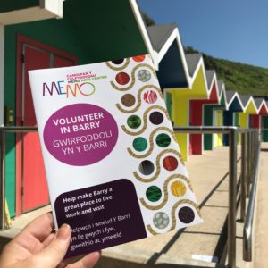A flyer is held in front of the colourful Barry Island beach huts on a gloriously sunny day. The flyer has the Memo logo and the words "Volunteer in Barry" in English and Welsh