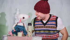 an actor in a tank top and bobble hat smiles at a puppet mouse over a flask