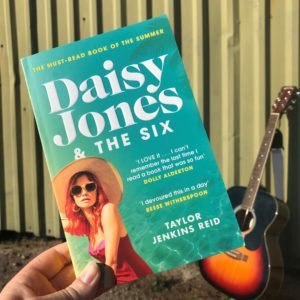 The book of Daisy Jones & The Six is held by a hand with a guitar in the background.