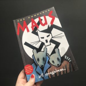 The book of Maus by Art Spiegelman is held against a dark background by a hand with chipped red nail varnish.