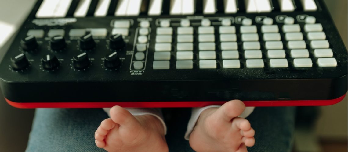 A piano keyboard sits on a baby's lap. The baby sits on an adult's lap. The baby's podgy little fingers are on the keys, their wriggly little feet poking out of the bottom in the foreground of the photo.