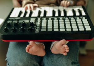 A piano keyboard sits on a baby's lap. The baby sits on an adult's lap. The baby's podgy little fingers are on the keys, their wriggly little feet poking out of the bottom in the foreground of the photo. 
