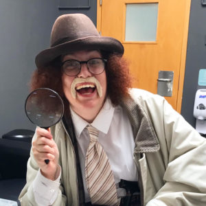 an actor in character with a magnifying glass and a fake moustache smiles at the camera