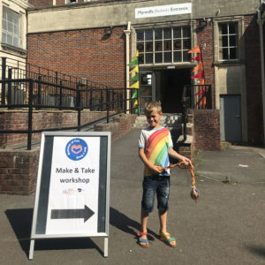 a boy in a rainbow tshirt swings his handmade poi next to a sign that reads Make and Take Workshop at the Box Office entrance to the Memo